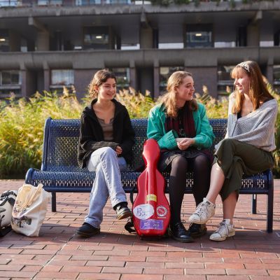 Three girls sit on a bench. From left to right: One wears a black cardigan, one wears a green jacket and the other wears a grey jumper. A red instrument case stands propped up against the bench in the middle of them.